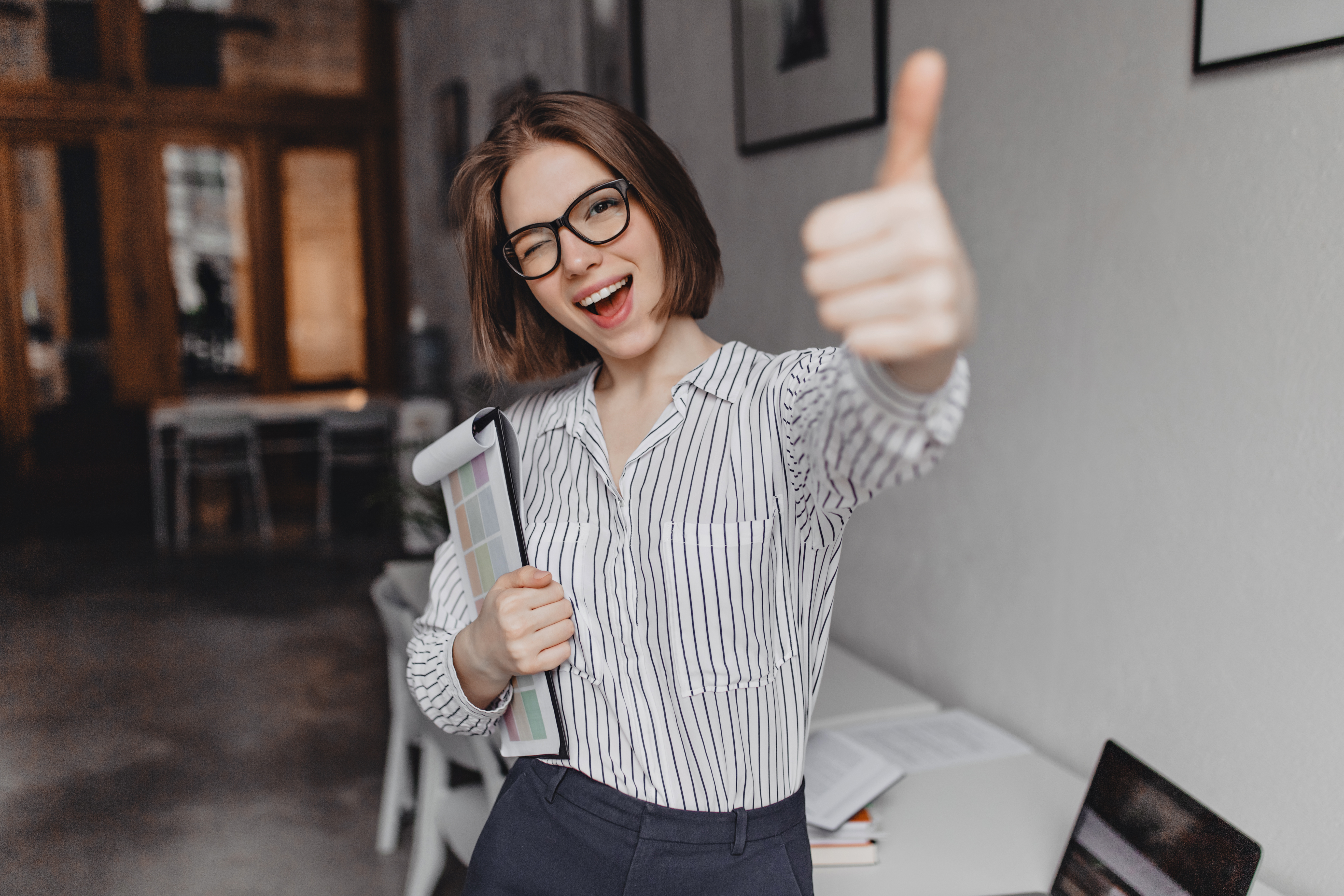 Young woman in office style clothes and glasses holds tablet with documents, winks and shows thumb up.
